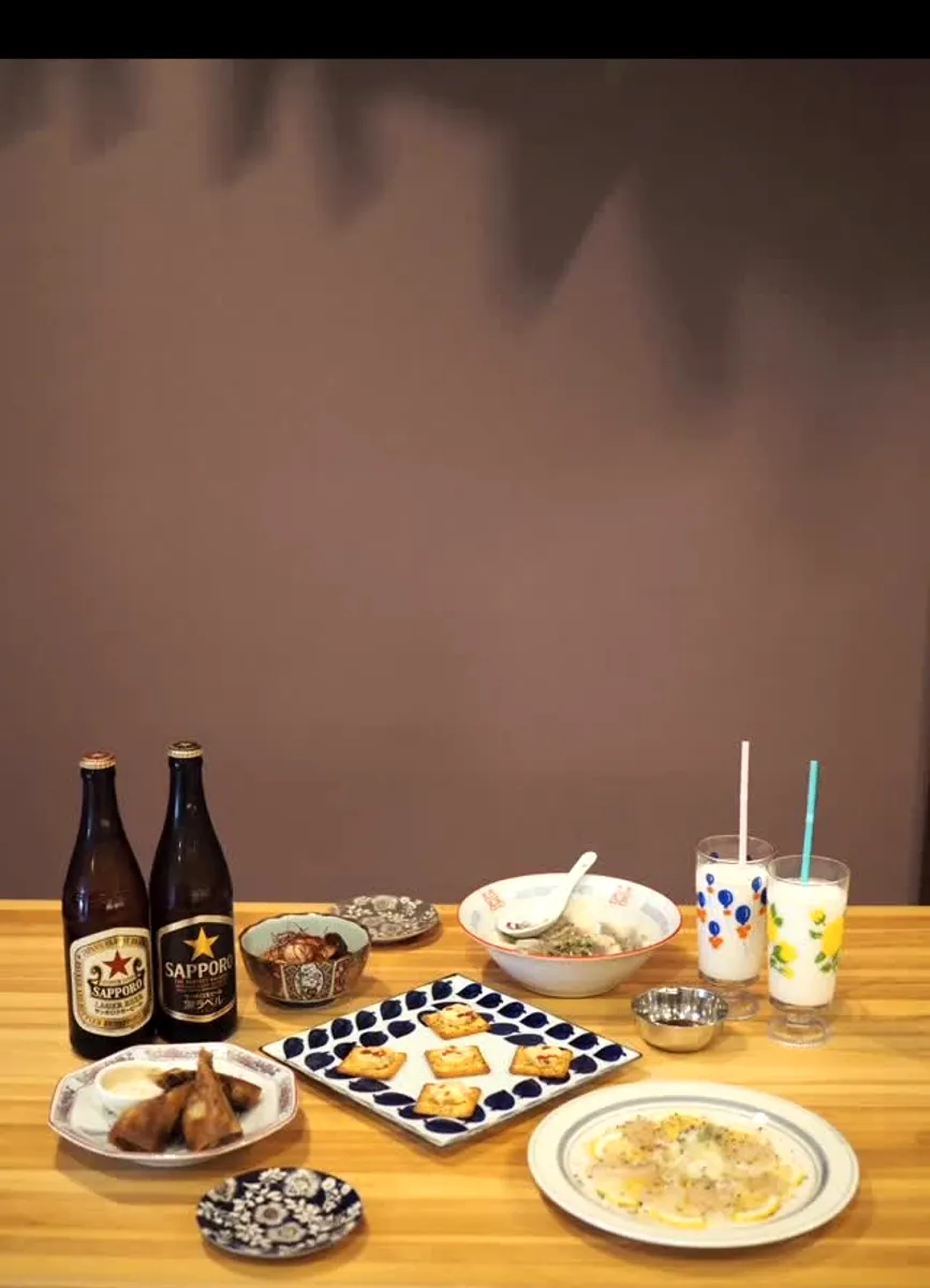 夜の卓上。瓶ビール2本と小皿のスパイス料理、クラッカーの盛り合わせ、ラッシーのグラス