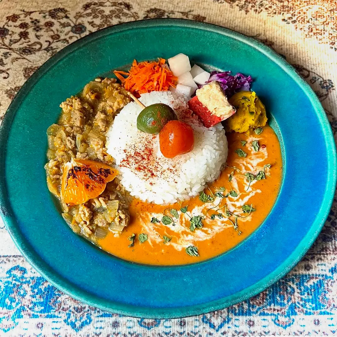 ターコイズブルーの大皿に、中央のライスを挟んでキーマカレーとオレンジ色のカレーが盛られたあいがけの一皿。ペルシャ絨毯柄の上で真俯瞰から